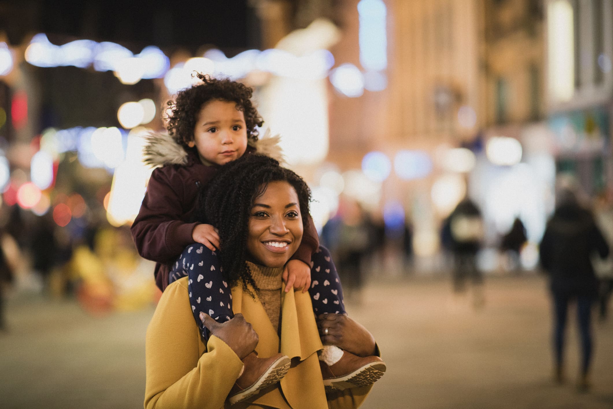 A close-up shot of a cute little girl sitting on her mother's shoulders, they are having a fun night together shopping for Christmas​ gifts.