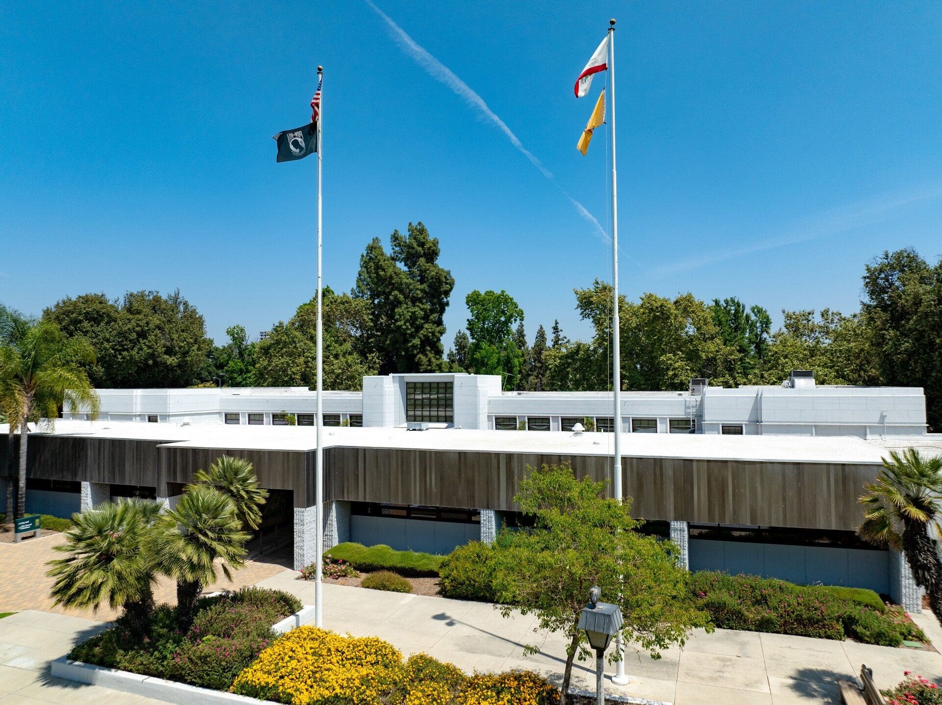 A modern, white-roofed building stands under a clear blue sky, flanked by trees. Two flags fly on tall poles, surrounded by lush greenery and colorful flowers.