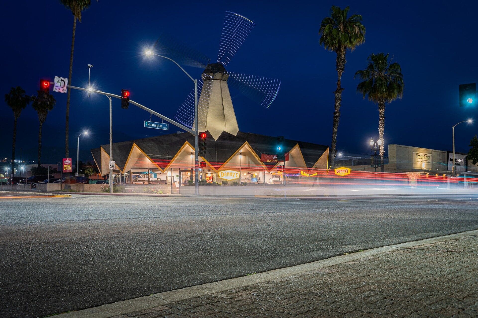 Nighttime scene of a diner with a windmill atop its roof, light trails from passing cars, palm trees, and traffic lights illuminating the urban setting.