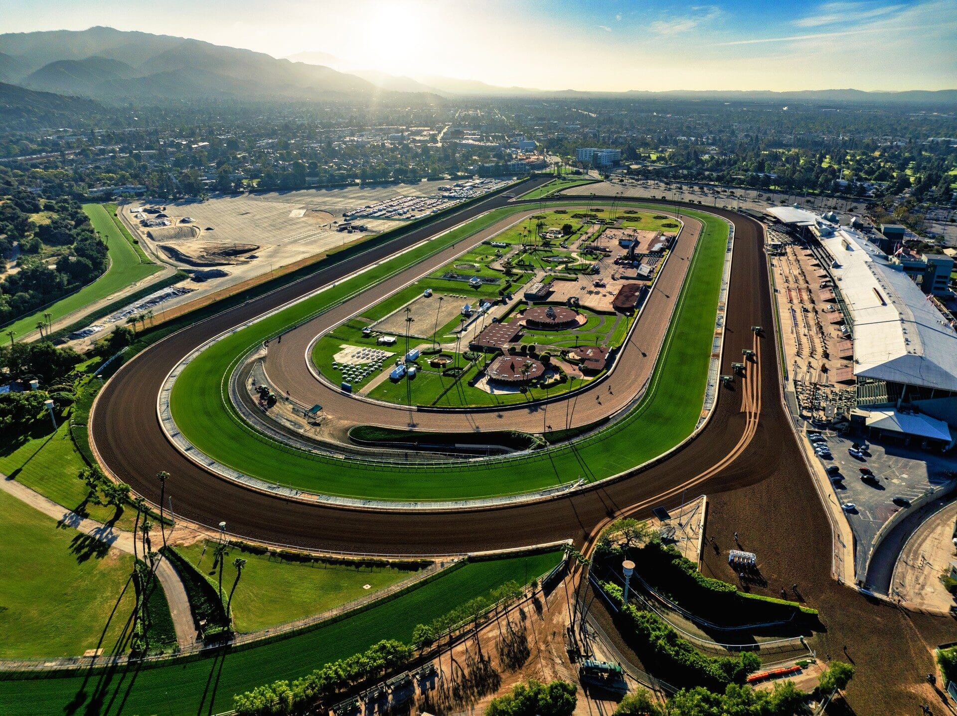 Aerial view of a sunlit horse racing track with expansive green fields and grandstands, set against a backdrop of mountains and a vast cityscape.