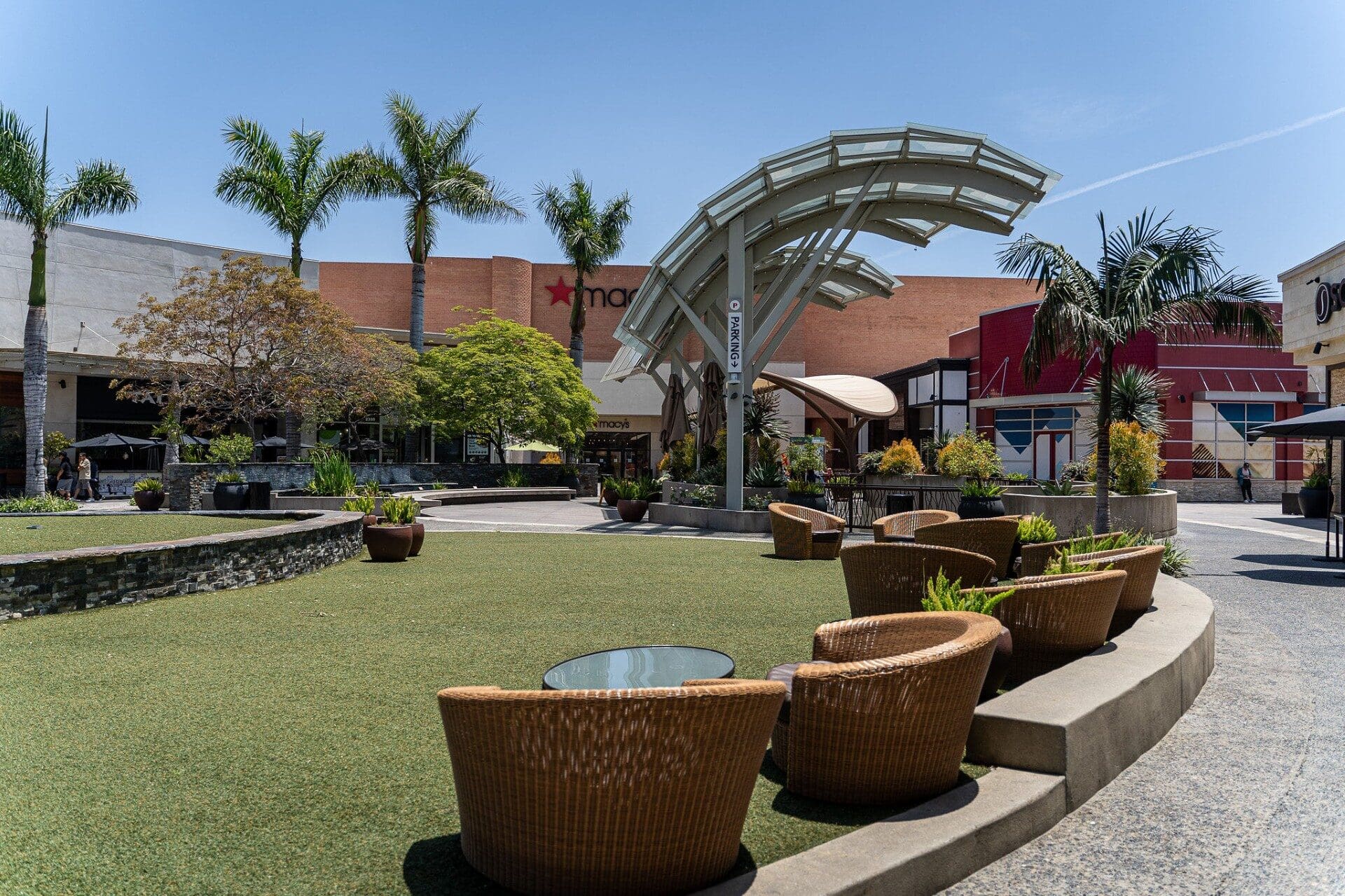 Outdoor mall scene with wicker chairs around a glass table on artificial grass. Palm trees and shops, including Macy's, create a relaxed atmosphere under a clear sky.