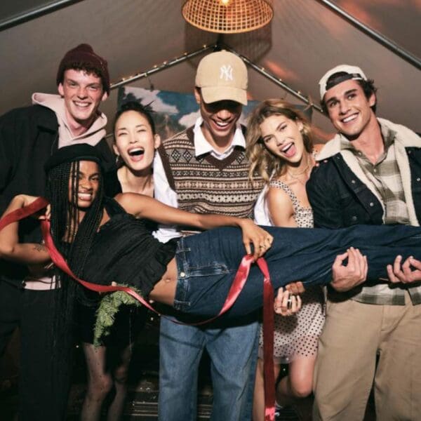 Six friends at a party pose joyfully with one friend being playfully lifted. They smile brightly, creating a lively and celebratory atmosphere.