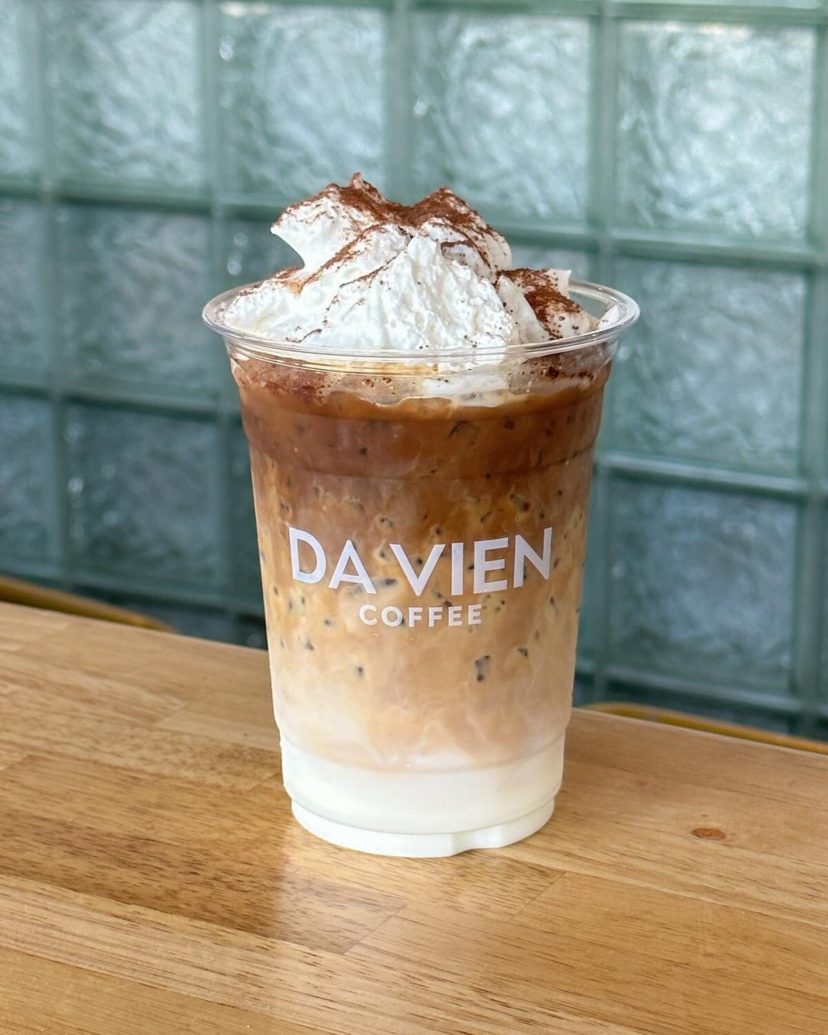 A creamy iced coffee from Da Vien Coffee with swirling layers of milk and espresso, topped with whipped cream and cocoa, on a wooden table.