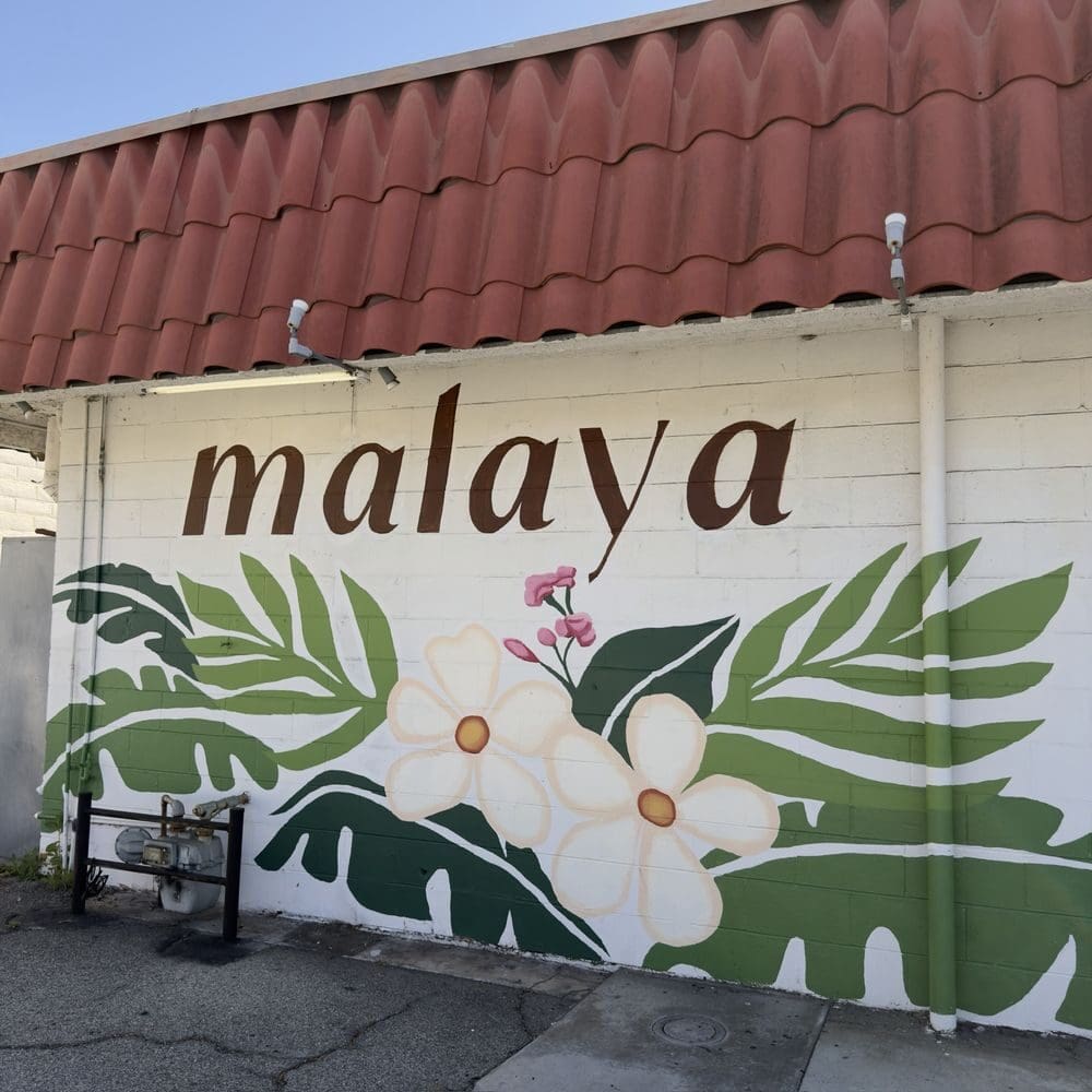 Mural on a white brick wall with the word "malaya" in brown, surrounded by large green leaves and white flowers. A serene and artistic vibe.